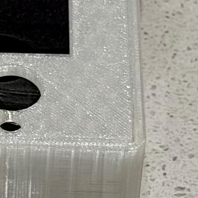 May include: A translucent, 3D-printed rectangular object with a textured surface finish. The piece features a large square cutout in the center and two circular holes positioned along the side, showing visible layer lines from the manufacturing process.