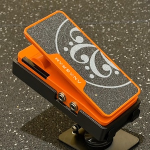 Puede incluir: Un pedal de expresión naranja y negro con una superficie de agarre negra texturizada, un gráfico de clave de fa en blanco y el texto ANAGRAM impreso en el lateral. El dispositivo incluye dos conectores de entrada circulares y un pedal abatible.