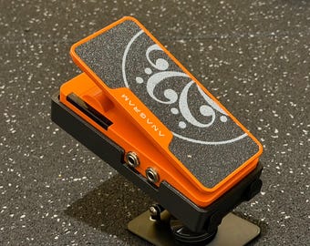 May include: An orange and black expression pedal featuring a textured black grip surface with a white bass clef graphic and the text ANAGRAM printed on the side. The device includes two circular input jacks on the side and a hinged foot treadle.