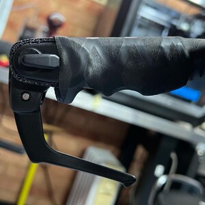 May include: A black bicycle brake lever featuring a textured, ergonomic grip with a repeating geometric pattern. The lever assembly includes a pivot point with a visible metal screw and a contoured handle designed for manual operation on a handlebar.