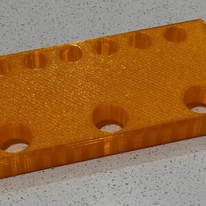 Puede incluir: Un bloque rectangular de pl&aacute;stico naranja con acabado brillante que presenta una fila de seis peque&ntilde;os orificios circulares en el borde superior y tres orificios circulares m&aacute;s grandes debajo, con una textura impresa en 3D en el lateral.