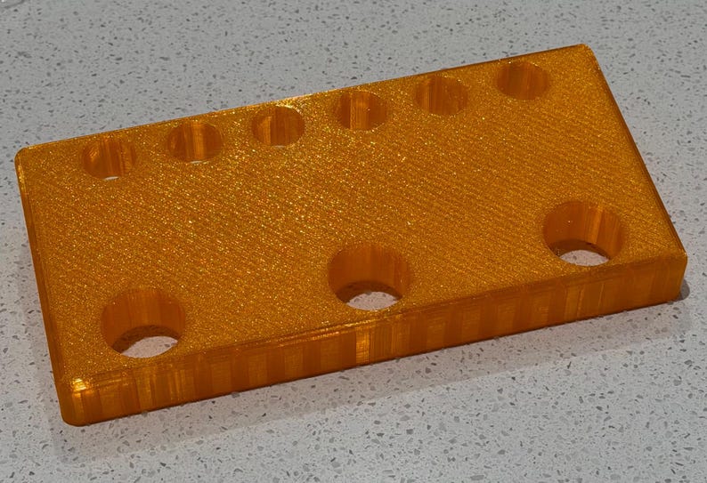 Puede incluir: Un bloque rectangular de pl&aacute;stico naranja con acabado brillante que presenta una fila de seis peque&ntilde;os orificios circulares en el borde superior y tres orificios circulares m&aacute;s grandes debajo, con una textura impresa en 3D en el lateral.