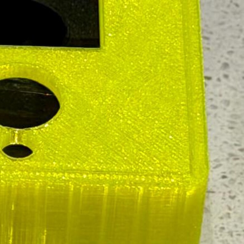 May include: A bright neon yellow 3D printed rectangular object with a textured surface. The object features a large square cutout at the top and a circular hole positioned below it, with visible layer lines throughout the structure.