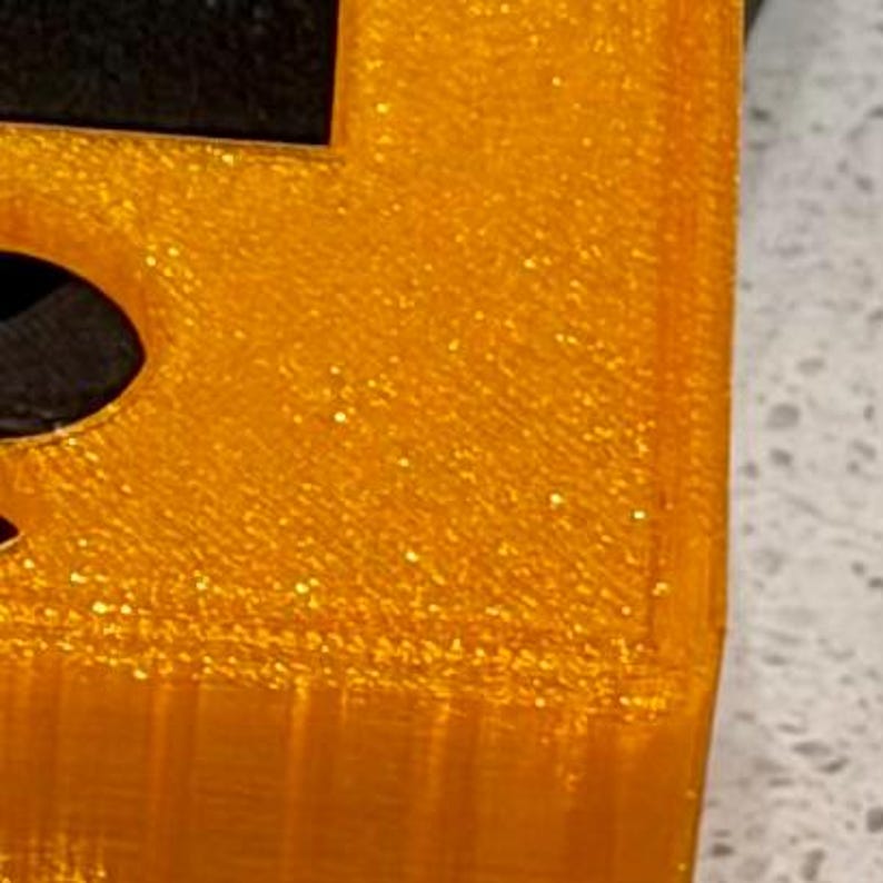 May include: A bright orange, glittery, three-dimensional printed object with a textured surface. The piece features a sharp corner and a flat top edge, showing visible layer lines from the manufacturing process and a distinct, sparkling finish.
