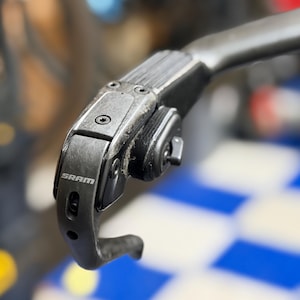 May include: A black SRAM bicycle brake lever and shifter component featuring a textured grip surface, a curved metal lever arm, and visible mounting screws. The brand name SRAM is printed in white lettering on the side of the black housing.