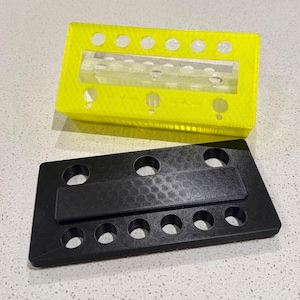Puede incluir: Un organizador rectangular de color amarillo ne&oacute;n impreso en 3D con un inserto central de acr&iacute;lico transparente y una serie de orificios circulares a lo largo de los bordes, junto a una base rectangular negra a juego con superficie texturizada.
