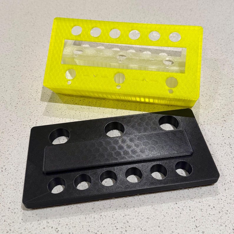 Puede incluir: Un organizador rectangular de color amarillo ne&oacute;n impreso en 3D con un inserto central de acr&iacute;lico transparente y una serie de orificios circulares a lo largo de los bordes, junto a una base rectangular negra a juego con superficie texturizada.