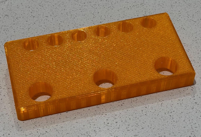 May include: A rectangular orange plastic block with a glittery finish featuring a row of six small circular holes along the top edge and three larger circular holes arranged in a row below, showing a layered 3D printed texture on the side.