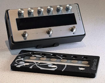 May include: A rectangular electronic device labeled ANAGRAM with a silver metallic top panel, six silver knobs, a black display screen, and three silver foot switches. A separate black plastic cover with white swirl patterns is placed in front.