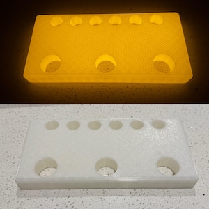 Puede incluir: Un soporte rectangular de pl&aacute;stico blanco con seis orificios circulares peque&ntilde;os a lo largo del borde superior y tres orificios circulares m&aacute;s grandes a lo largo del borde inferior. El objeto se muestra en dos estados: brillante y blanco.
