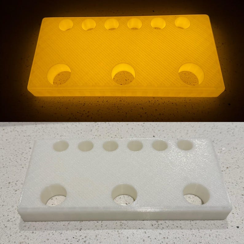 Puede incluir: Un soporte rectangular de pl&aacute;stico blanco con seis orificios circulares peque&ntilde;os a lo largo del borde superior y tres orificios circulares m&aacute;s grandes a lo largo del borde inferior. El objeto se muestra en dos estados: brillante y blanco.