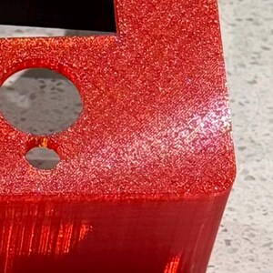 May include: A bright red, glittery, three-dimensional object with a textured surface. The object features a large circular opening positioned above a smaller circular opening, with sharp, defined edges and a distinct, granular, sparkling finish.