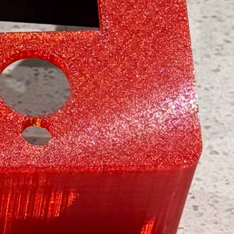 May include: A bright red, glittery, three-dimensional object with a textured surface. The object features a large circular opening positioned above a smaller circular opening, with sharp, defined edges and a distinct, granular, sparkling finish.