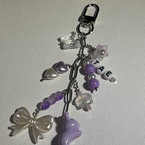 May include: A silver-toned metal keychain featuring a variety of charms including a white pearlescent bow, a light purple rabbit, a clear butterfly, a seashell, heart-shaped beads, and letter beads spelling out the name TAE in black on white.