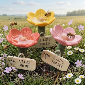 May include: Three flower-shaped water stations for bees in yellow, coral, and pink. Each features a central textured disc, a green stem, and a rectangular sign reading CUPS FOR BEE with a small bee icon. Several bees are visible on the petals.