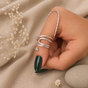 Könnte beinhalten: Ein silberfarbener Fingerpanzerring mit einem langen, geschwungenen vertikalen Stab, der sich entlang des Fingers erstreckt, verbunden mit einer Reihe von drei horizontalen, gestapelten, offenen Bändern, die den Finger umschließen, mit einem kleinen Vier-Punkt-Akzent.