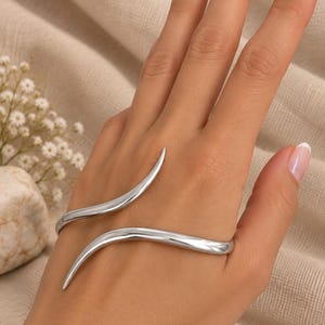 Könnte beinhalten: Ein poliertes Handarmband in Silberoptik mit einem eleganten, offenen Design, das aus zwei geschwungenen, spitz zulaufenden Armen besteht, die sich über den Handrücken legen. Das Metall hat eine glatte, reflektierende Oberfläche und Form.