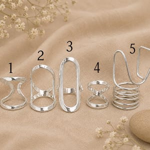 May include: Five silver-colored metal finger splints labeled with numbers one through five. Ring one is a curved band, two is a tall oval, three is an elongated oval, four is a crisscross band, and five features a coiled base with a tall wire loop.