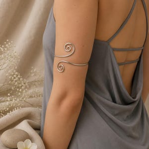 May include: A silver-toned metal arm cuff featuring a continuous wire design that forms two symmetrical spirals at the ends. The arm cuff is worn on the upper arm, showcasing a smooth, polished finish that wraps around the bicep area.