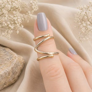 Könnte beinhalten: Ein goldfarbener Metallring mit einem durchgehenden, spiralförmigen Design, das sich um den Finger wickelt. Das Band besteht aus einem einzelnen Draht, der ein Zickzackmuster bildet und an beiden Enden in kleinen, abgerundeten Kugeln endet.