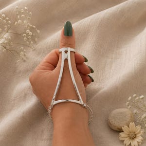 Könnte beinhalten: Eine silberne Handkette mit einem dreieckigen Metallrahmen, der einen am Daumen getragenen Ring mit einem zarten Kettenarmband um das Handgelenk verbindet. Das polierte Metallstück weist ein geometrisches Design mit einer Aussparung auf.