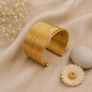 Könnte beinhalten: Ein breites, goldfarbenes Armband, das aus mehreren dünnen, parallelen Drahtreifen besteht. Das Design zeigt eine Reihe verbundener, kreisförmiger Drähte, die eine starre, offene Struktur bilden und an beiden Enden mit kleinen Kugeln abschließen.