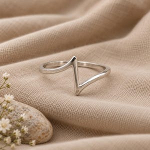 May include: A polished silver ring featuring a distinct zigzag or heartbeat-shaped focal point. The band is smooth and continuous, forming a sharp, angular peak in the center that rises above the circular profile of the ring's main body.