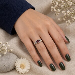 Könnte beinhalten: Ein Silberring mit einem tiefroten Granat im Marquise-Schliff, gefasst in ein dekoratives Filigranband. Der Ring wird an einem Finger mit dunkelgrünem Nagellack getragen und zeigt die filigranen Metallarbeiten um den Stein.