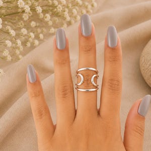 Könnte beinhalten: Ein silberfarbener Ring mit einem breiten Design, bestehend aus zwei parallelen horizontalen Stäben oben und unten, die durch zwei vertikale, geschwungene Segmente an jeder Seite verbunden sind und eine offene, geometrische Struktur bilden.