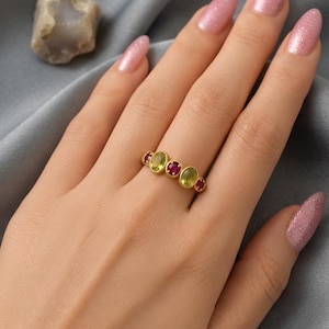 Könnte beinhalten: Ein goldener Ring mit einer Reihe von fünf abwechselnd oval geschliffenen Edelsteinen, bestehend aus drei hellgrünen Peridoten und zwei dunkelroten Rubinen, die in einem einfachen Metallband gefasst sind und horizontal auf dem Finger liegen.