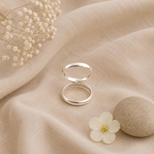 May include: A silver ring featuring a double band design with two parallel circular loops connected by a vertical bar. The ring has a smooth, polished metallic finish and is positioned upright on a soft, light beige fabric surface.
