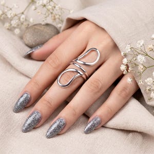 Könnte beinhalten: Ein silberfarbener Ring mit einem länglichen Design aus offenen Schlaufen, das sich über zwei Fingergelenke erstreckt. Das Metall hat eine glatte, polierte Oberfläche und die Struktur besteht aus zwei verbundenen, tropfenförmigen Schlaufen.