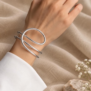 Könnte beinhalten: Ein silberfarbenes Armband mit einem fließenden, mehrschichtigen Drahtdesign, das sich um das Handgelenk legt. Das Armband hat eine glatte, polierte metallische Oberfläche und eine offene, überlappende Struktur, die eine kreisförmige Schlaufe bildet.