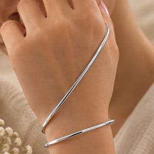 Könnte beinhalten: Ein Handarmband aus poliertem, silberfarbenem Metall mit einem durchgehenden, glatten und schlanken Band, das sich diagonal vom Handgelenk über den Handrücken windet und eine elegante, minimalistische und reflektierende Kurvenform bildet.