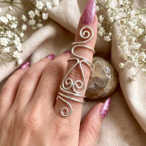 Könnte beinhalten: Ein silberfarbener Drahtring mit einem länglichen Design, das einen großen Teil des Fingers bedeckt. Der Ring besteht aus einem einzigen durchgehenden Stück Metalldraht, das zu mehreren Spiralen, Schlaufen und herzähnlichen Mustern geformt ist.