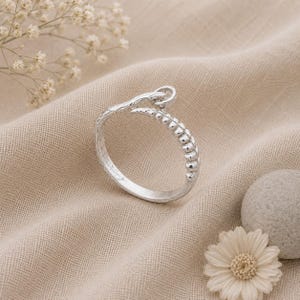 Könnte beinhalten: Ein silberfarbener Ring mit offenem Design, der auf einer Seite eine texturierte, perlenartige Oberfläche und auf der anderen Seite ein glattes, zulaufendes Ende aufweist. Das Band biegt sich oben zu einer kleinen Schlaufe.