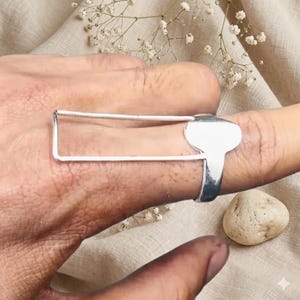 Könnte beinhalten: Ein silberfarbener Ring mit einem markanten rechteckigen Rahmendesign, das sich horizontal über den Finger erstreckt. Die Ringschiene ist glatt und poliert und mit einer massiven, abgerundeten Metallplatte verbunden, die flach am Finger anliegt.