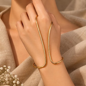 Könnte beinhalten: Ein Handarmband aus goldfarbenem Metall mit einem glatten, durchgehenden Drahtdesign, das sich um das Handgelenk legt und nach oben führt, um einen Finger zu umschließen, wodurch eine offene, geschwungene Silhouette auf dem Handrücken entsteht.