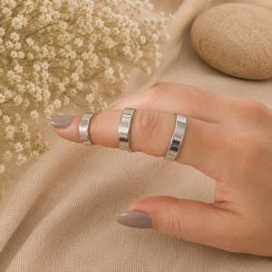 May include: Three polished silver-tone metal rings with a smooth, flat band design. Each ring features a consistent width and a reflective, metallic finish. The rings are worn on a finger, showcasing their simple, unadorned, and uniform appearance.