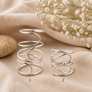 Könnte beinhalten: Zwei Ringe aus silberfarbenem Metall. Ring 1 hat ein hohes, mehrschichtiges Spiraldesign. Ring 2 hat ein herzförmiges Drahtdesign, das an den Enden und Verbindungsstellen mit kleinen kugelförmigen Perlen verziert ist. Beide Ringe sind mit 1 und 2 markiert.