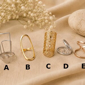 Könnte beinhalten: Fünf verschiedene Metallringe, beschriftet mit A bis E. Ring A ist ein silberner rechteckiger Rahmen, B ein glatter goldener Ovalring, C ein goldener filigraner Zylinder, D ein silbernes Design mit geteiltem Band und E ein roségoldener offener Ring.