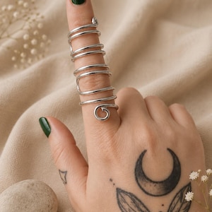 Könnte beinhalten: Ein Ring aus silberfarbenem Metall mit einem durchgehenden Spiraldesign, das sich mehrfach um den Finger wickelt und von der Basis bis zur Spitze reicht. Der Ring hat eine polierte, glatte Oberfläche und ein kreisförmiges Spulendetail am unteren Ende.