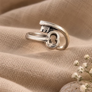 Könnte beinhalten: Ein Ring aus silberfarbenem Metall in Form eines antiken Schlüssels. Die Ringschiene biegt sich zur Form des Schlüsselkopfes, während der Schlüsselbart horizontal über die Oberseite verläuft. Das Metall hat ein leicht oxidiertes Finish.