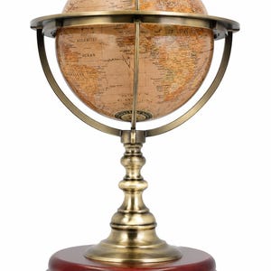 Puede incluir: Un globo terráqueo con un mapa en tonos sepia y texto en negro, montado sobre un anillo meridiano de metal color latón y un soporte decorativo. El conjunto descansa sobre una base circular de madera pulida en color marrón rojizo.