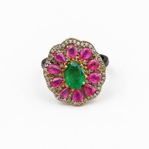May include: A ring featuring a central oval-cut green emerald surrounded by a halo of pear-shaped pink rubies. The floral-inspired setting is accented with small white pavé diamonds set in gold, mounted on a smooth, solid black band.