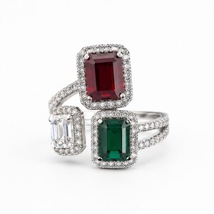 Könnte beinhalten: Ein Bypass-Ring mit drei Steinen im Smaragdschliff: ein tiefroter Rubin, ein leuchtend grüner Smaragd und ein klarer weißer Diamant. Jeder Stein ist von einem Kranz aus kleinen runden Pavé-Diamanten umgeben, gefasst in eine silberfarbene Ringschiene.