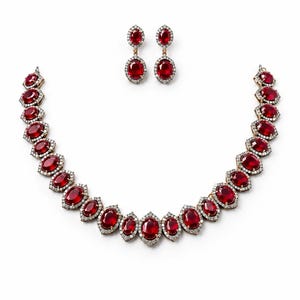 Puede incluir: Un conjunto de joyería compuesto por un collar y un par de pendientes colgantes. Cada pieza presenta piedras de color rojo intenso en forma ovalada rodeadas por un borde de pequeñas piedras blancas claras en un marco metálico.