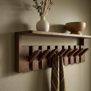 May include: A wall-mounted wooden coat rack featuring a horizontal shelf and multiple angled hooks. A white ceramic vase with dried foliage and a wooden bowl sit on the shelf. A patterned olive green scarf hangs from one of the wooden hooks.