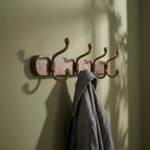 May include: A wall-mounted coat rack featuring a horizontal wooden base with a natural grain pattern and four curved brass-toned metal hooks. A grey fabric garment hangs from one of the hooks, partially obscuring the lower portion of the rack.