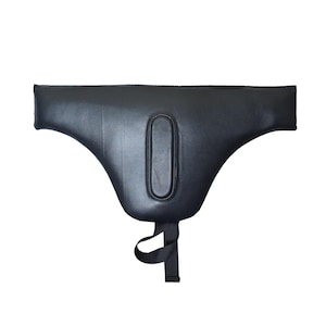 May include: A black protective groin guard featuring a smooth, contoured surface with a central oval-shaped reinforcement panel. The piece includes a black fabric strap extending from the bottom center, designed for secure attachment and support.